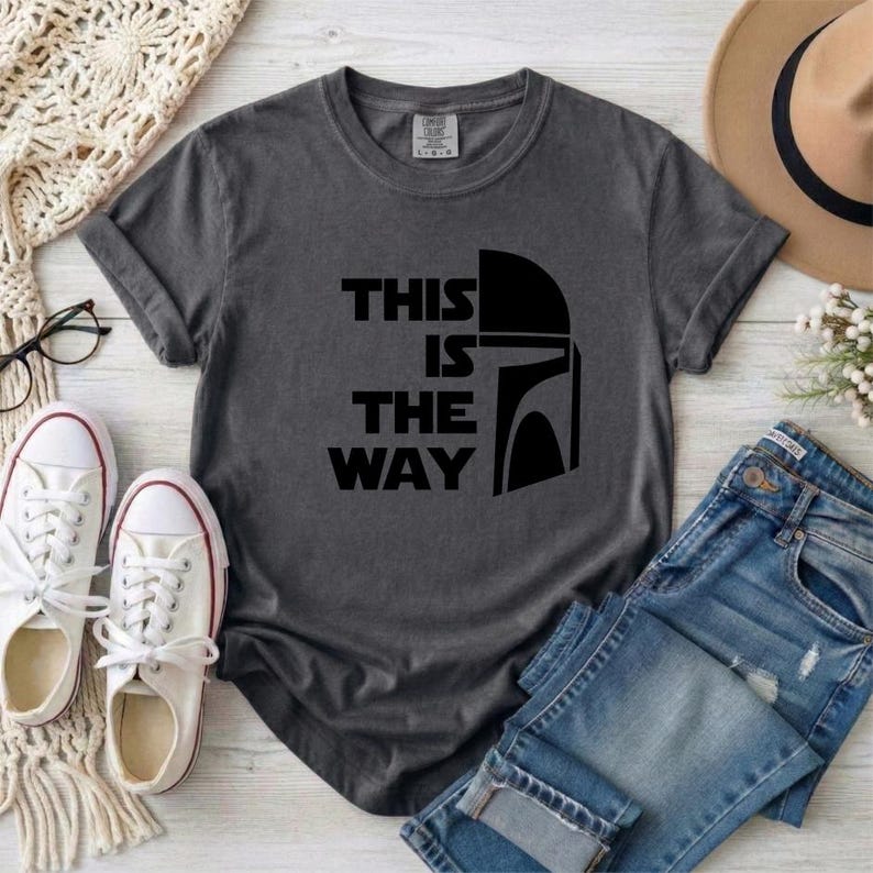 May include: Dark gray t-shirt with the text "THIS IS THE WAY" and a black helmet graphic. The shirt is paired with blue jeans and white sneakers. A tan hat and a cream-colored scarf are also in the image.