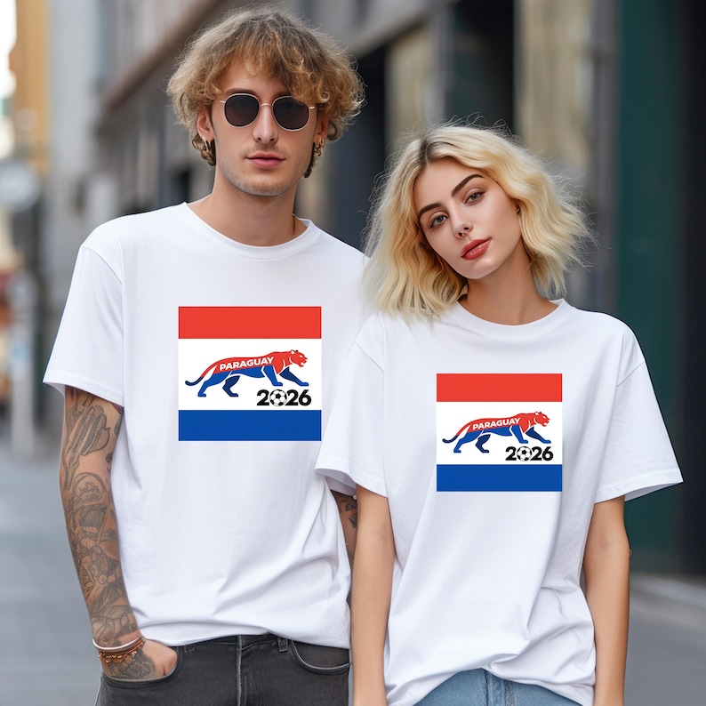 Pu&ograve; includere: Due persone indossano magliette bianche. Ogni maglietta presenta un design rettangolare con strisce rosse, bianche e blu, una tigre blu, la parola "PARAGUAY" e il numero "2026".