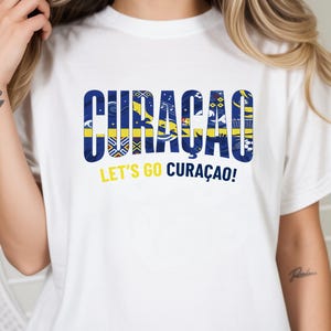 May include: White t-shirt with the word "CURACAO" in blue and yellow, featuring various patterns within the letters. Below the word is the phrase "LET'S GO CURA&Ccedil;AO!" in yellow. The shirt is a casual wear item.