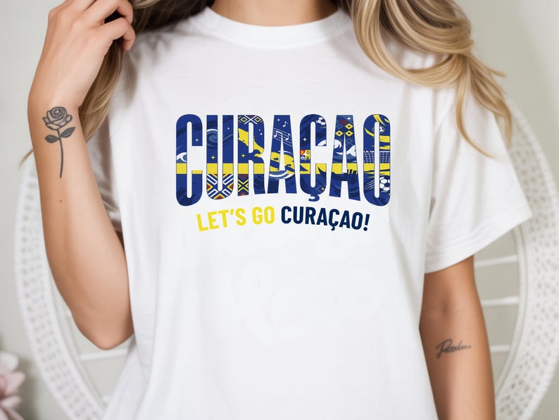 May include: White t-shirt with the word "CURACAO" in blue and yellow, featuring various patterns within the letters. Below the word is the phrase "LET'S GO CURA&Ccedil;AO!" in yellow. The shirt is a casual wear item.