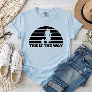 May include: Light blue t-shirt with a black graphic of a silhouette of a figure and a child against a striped background. The text "THIS IS THE WAY" is below the graphic. The shirt is displayed with white sneakers and blue jeans.