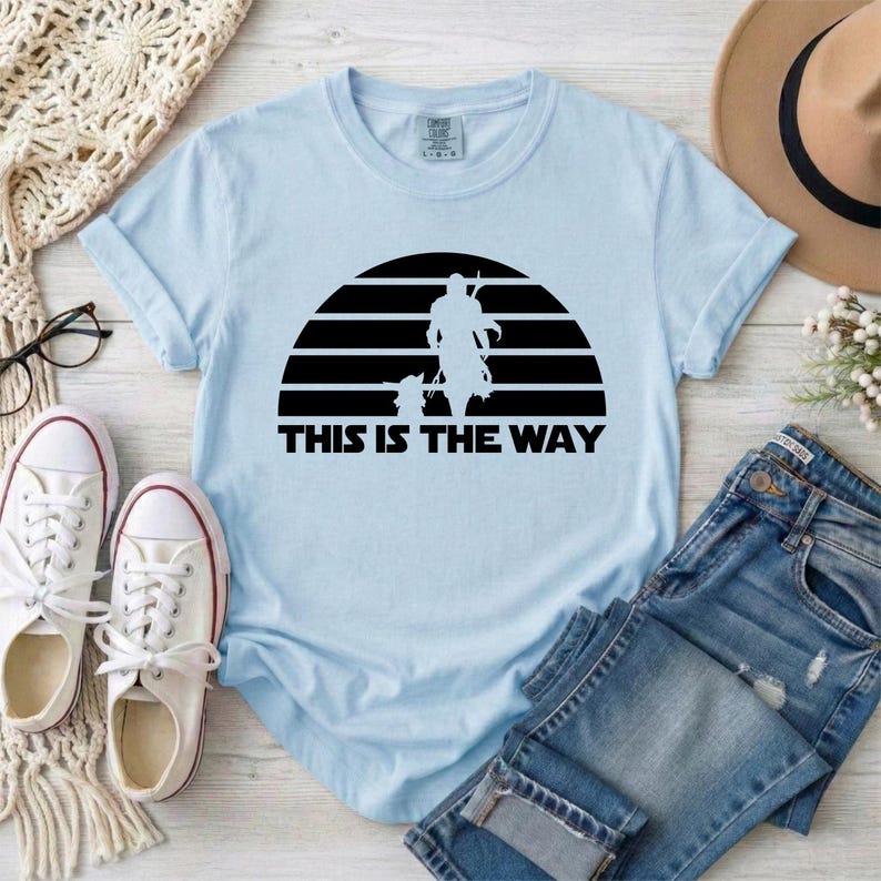 May include: Light blue t-shirt with a black graphic of a silhouette of a figure and a child against a striped background. The text "THIS IS THE WAY" is below the graphic. The shirt is displayed with white sneakers and blue jeans.