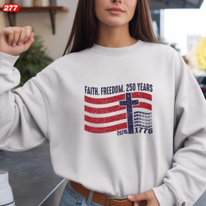 Pode incluir: Camisola branca com um gr&aacute;fico desgastado de uma bandeira americana, uma cruz e o texto "Faith. Freedom. 250 Years". O design utiliza vermelho e azul, com os anos 1776 e 2026. A camisola &eacute; de estilo casual.