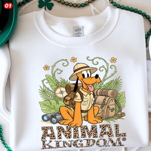 May include: White sweatshirt featuring a cartoon Pluto character in safari attire, with a backpack, binoculars, and the words "ANIMAL KINGDOM" in leopard print lettering. The design includes green foliage and a compass.