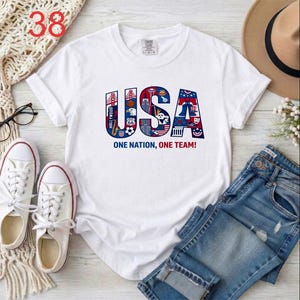 Puede incluir: Una camiseta blanca de manga corta con un gr&aacute;fico grande de las letras USA en rojo, blanco y azul. Las letras est&aacute;n rellenas con varios iconos y monumentos estadounidenses. Debajo del gr&aacute;fico, el texto dice ONE NATION, ONE TEAM!