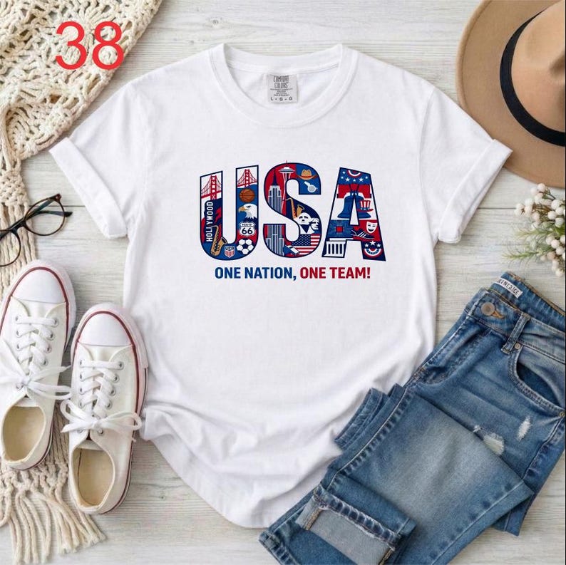 Puede incluir: Una camiseta blanca de manga corta con un gr&aacute;fico grande de las letras USA en rojo, blanco y azul. Las letras est&aacute;n rellenas con varios iconos y monumentos estadounidenses. Debajo del gr&aacute;fico, el texto dice ONE NATION, ONE TEAM!