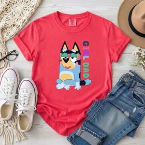 May include: A coral red short-sleeve t-shirt featuring a graphic of a blue and yellow cartoon dog sitting with its legs crossed. The text "GIRL DAD" is printed vertically in colorful letters next to the dog. The shirt has a crew neckline.