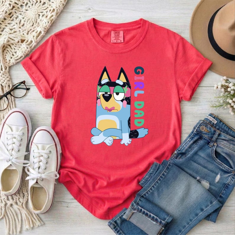 May include: A coral red short-sleeve t-shirt featuring a graphic of a blue and yellow cartoon dog sitting with its legs crossed. The text "GIRL DAD" is printed vertically in colorful letters next to the dog. The shirt has a crew neckline.
