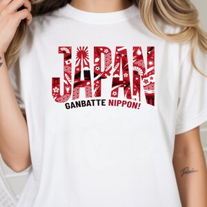 May include: White t-shirt with the word "JAPAN" in red, stylized with Japanese-themed illustrations. Below, the phrase "GANBATTE NIPPON!" is printed in black. The shirt is a casual wear item.