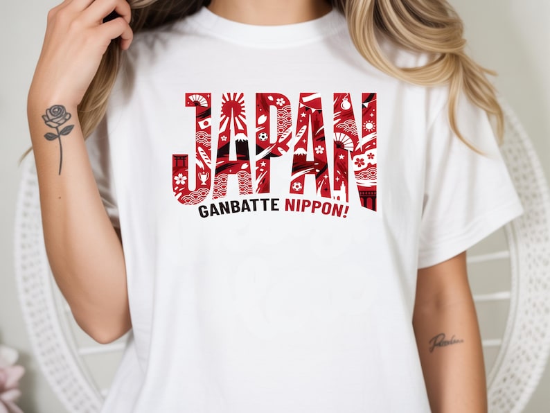 May include: White t-shirt with the word "JAPAN" in red, stylized with Japanese-themed illustrations. Below, the phrase "GANBATTE NIPPON!" is printed in black. The shirt is a casual wear item.