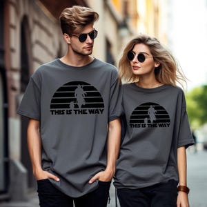 May include: Two people wearing matching gray t-shirts with a black graphic design. The design features a silhouette of a figure and a creature within a half-circle, with the text "THIS IS THE WAY" below. Both are wearing sunglasses.