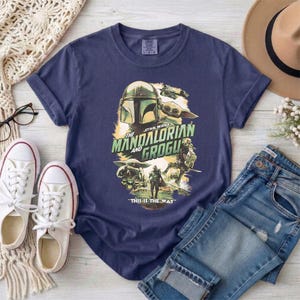May include: Navy blue t-shirt featuring a graphic from the Star Wars series, "The Mandalorian and Grogu." The design includes images of the Mandalorian, Grogu, and other characters. The text "This is the Way" is also included in the design.