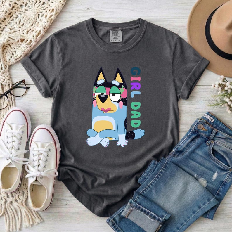 May include: A charcoal gray short-sleeve t-shirt featuring a graphic of a blue cartoon dog with a pink headband and a yellow belly. The text "GIRL DAD" is printed vertically in colorful block letters next to the dog. The shirt has a crew neckline.