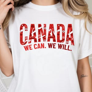 May include: White t-shirt with the word "CANADA" in large red letters filled with sports and winter-themed graphics. Below the word "CANADA" is the text "WE CAN. WE WILL."
