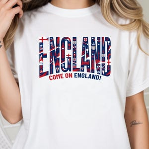 May include: White t-shirt with the word "ENGLAND" in large, stylized letters. The letters are filled with red and blue patterns and symbols. Below the word is the phrase "COME ON ENGLAND!"