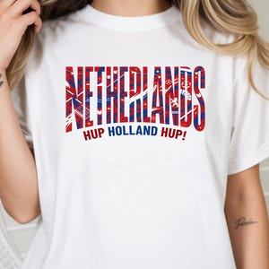 May include: White t-shirt with the word "NETHERLANDS" in red and blue block letters. Below the word is the phrase "HUP HOLLAND HUP!" in red. The shirt has short sleeves.