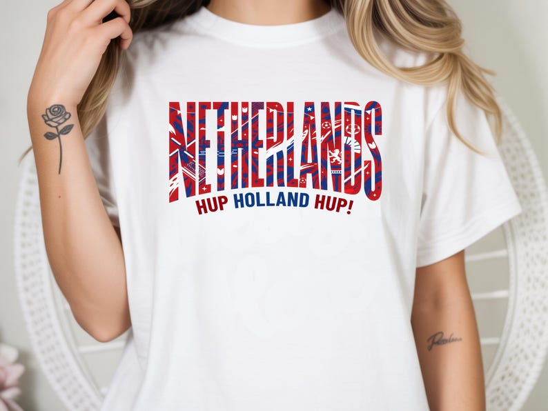 May include: White t-shirt with the word "NETHERLANDS" in red and blue block letters. Below the word is the phrase "HUP HOLLAND HUP!" in red. The shirt has short sleeves.