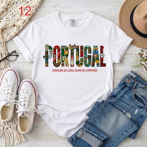 May include: White t-shirt with the word "PORTUGAL" in colorful, decorative letters. Below the word is the text "CORA&Ccedil;&Atilde;O DE LE&Atilde;O, ALMA DE CAMPE&Atilde;O". The shirt is paired with blue jeans, white sneakers, and a hat.