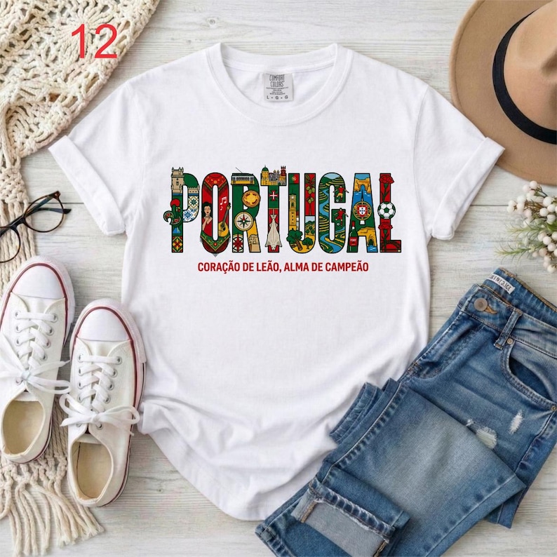 May include: White t-shirt with the word "PORTUGAL" in colorful, decorative letters. Below the word is the text "CORA&Ccedil;&Atilde;O DE LE&Atilde;O, ALMA DE CAMPE&Atilde;O". The shirt is paired with blue jeans, white sneakers, and a hat.