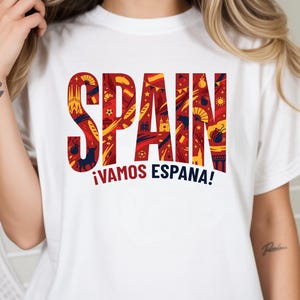 May include: White t-shirt with the word "SPAIN" in large, red letters filled with yellow and blue illustrations. Below, the phrase "&iexcl;VAMOS ESPA&Ntilde;A!" is in red and blue. The shirt is a casual wear item.