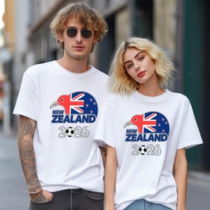 Pu&ograve; includere: Due persone indossano magliette bianche. Ogni maglietta presenta un disegno con un kiwi, la bandiera della Nuova Zelanda e la scritta "NEW ZEALAND 2026" con un pallone da calcio. Il design &egrave; in rosso, blu e bianco.