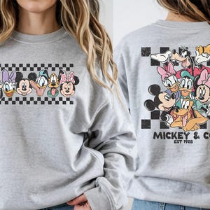 Pode incluir: Um moletom cinza claro com um gr&aacute;fico colorido de personagens da Disney. A frente exibe rostos de personagens acima de um padr&atilde;o xadrez. A parte de tr&aacute;s mostra um grupo de personagens com o texto "Mickey & Co. Est 1928".