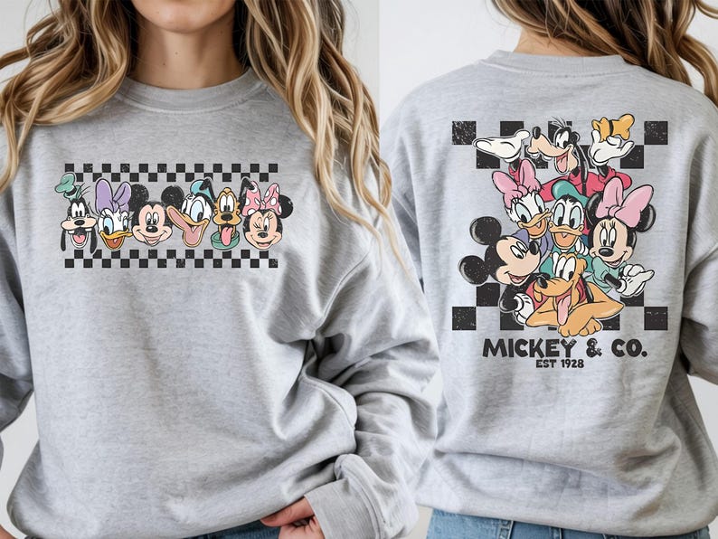 Pode incluir: Um moletom cinza claro com um gr&aacute;fico colorido de personagens da Disney. A frente exibe rostos de personagens acima de um padr&atilde;o xadrez. A parte de tr&aacute;s mostra um grupo de personagens com o texto "Mickey & Co. Est 1928".