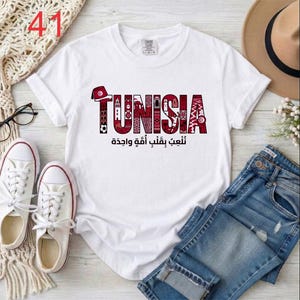 Puede incluir: Una camiseta blanca de manga corta con la palabra TUNISIA en letras rojas grandes rellenas de patrones intrincados e iconos culturales. Debajo del texto, la escritura &aacute;rabe dice نلعب بقلب أمة واحدة. El dise&ntilde;o incluye un icono de un fez rojo.