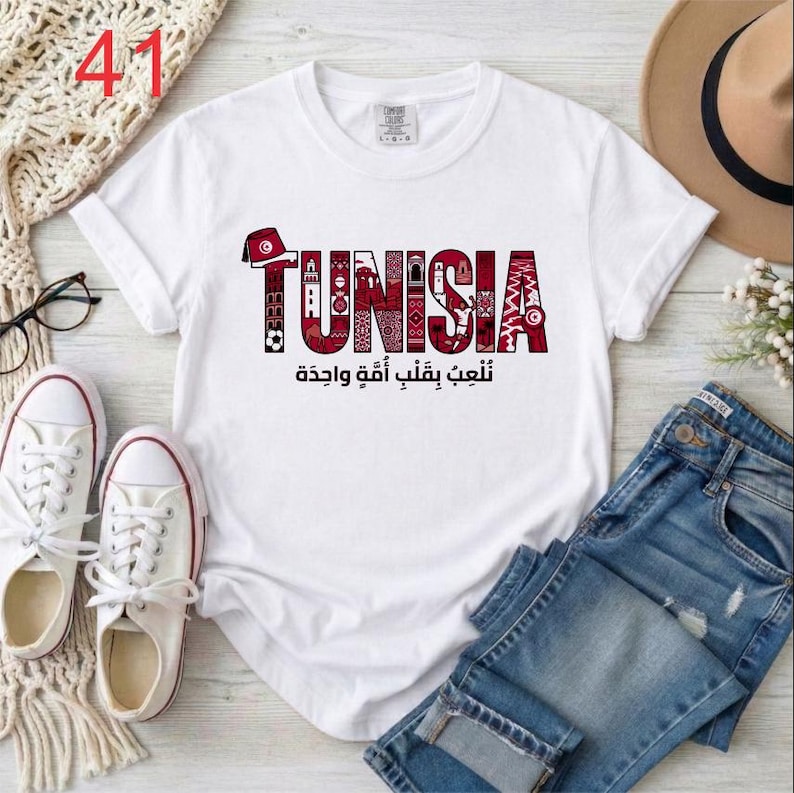 Puede incluir: Una camiseta blanca de manga corta con la palabra TUNISIA en letras rojas grandes rellenas de patrones intrincados e iconos culturales. Debajo del texto, la escritura &aacute;rabe dice نلعب بقلب أمة واحدة. El dise&ntilde;o incluye un icono de un fez rojo.