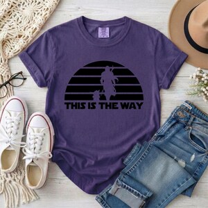 May include: A purple t-shirt with a black graphic of a figure and child against a sunset design. The text "THIS IS THE WAY" is below the graphic. The shirt is paired with blue jeans and white sneakers.