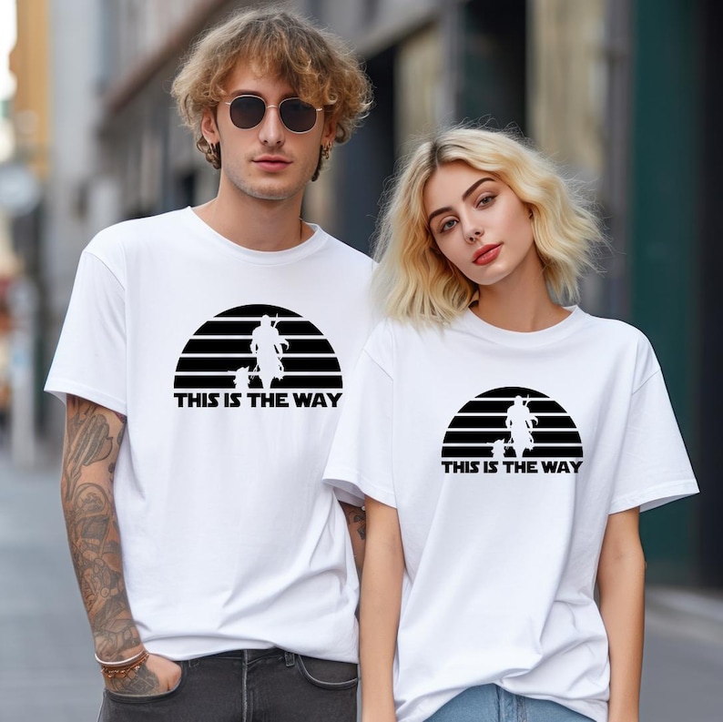 May include: White t-shirts featuring a black graphic design of a figure and the text "THIS IS THE WAY" inside a black semi-circle. The shirts are short-sleeved and worn by two people. The background is a blurred outdoor setting.