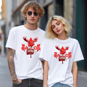 May include: Two people wearing white t-shirts with a red graphic featuring a moose head, the word "CANADA", and the year "2026" on a maple leaf design. The shirts are short-sleeved.
