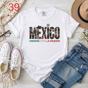 Puede incluir: Una camiseta blanca de manga corta que presenta la palabra MEXICO en letras grandes y estampadas rellenas de motivos culturales en verde, rojo y negro, con el texto UNIDOS POR LA PASI&Oacute;N impreso en letras m&aacute;s peque&ntilde;as de color verde y rojo debajo.