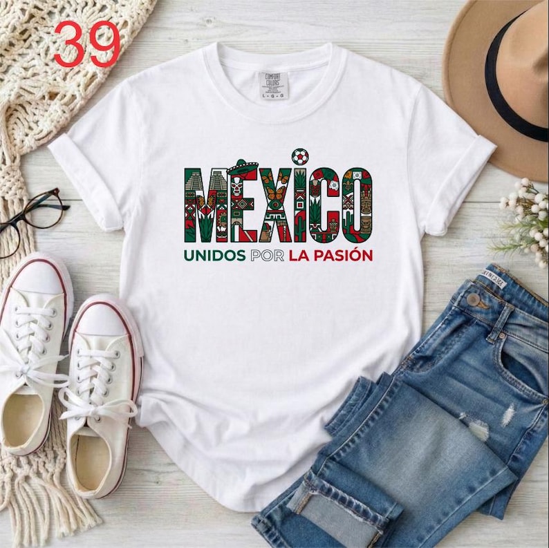 Puede incluir: Una camiseta blanca de manga corta que presenta la palabra MEXICO en letras grandes y estampadas rellenas de motivos culturales en verde, rojo y negro, con el texto UNIDOS POR LA PASI&Oacute;N impreso en letras m&aacute;s peque&ntilde;as de color verde y rojo debajo.