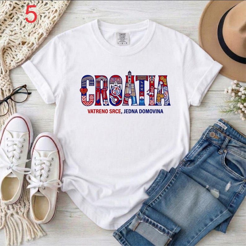May include: White t-shirt with the word "CROATIA" in a colorful, patterned design. Below the word is the text "VATRENO SRCE, JEDNA DOMOVINA". The shirt is paired with blue jeans and white sneakers.