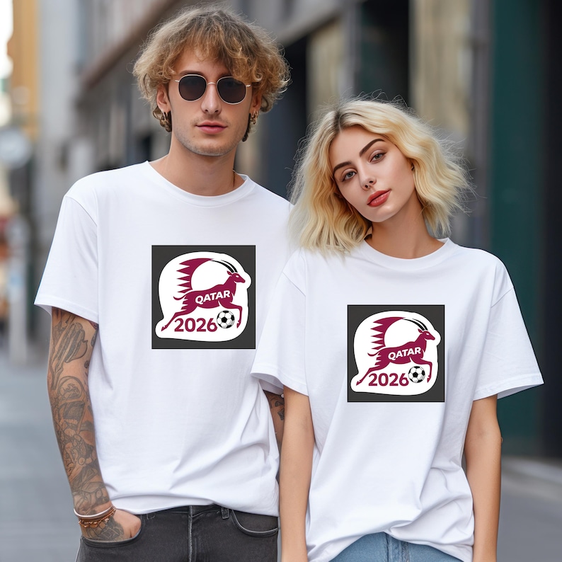 Pu&ograve; includere: Due persone indossano magliette bianche. Ogni maglietta presenta una grafica bordeaux e bianca di una capra che corre, un pallone da calcio e la scritta "QATAR 2026" in un quadrato nero.