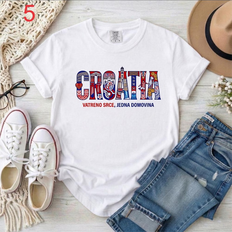 May include: White t-shirt featuring the word "CROATIA" in a vibrant, patterned design. The text "VATRENO SRCE, JEDNA DOMOVINA" is below. The shirt is styled with blue denim jeans and white trainers.