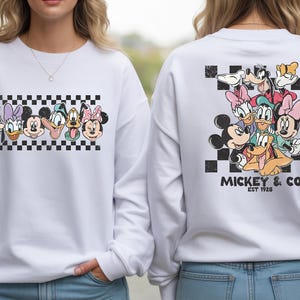 Pode incluir: Camisola branca com um gr&aacute;fico do Mickey Mouse e seus amigos. O design frontal apresenta rostos de personagens em um padr&atilde;o xadrez preto e branco. O design traseiro apresenta um grupo de personagens com o texto "Mickey & Co. EST 1928".