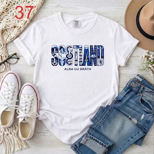 Puede incluir: Una camiseta blanca de manga corta con la palabra SCOTLAND en letras grandes de bloque con patrones azules y blancos, que contienen varios iconos escoceses. Debajo de la palabra, el texto ALBA GU BR&Agrave;TH est&aacute; impreso en azul oscuro.