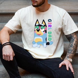 May include: A cream-colored short-sleeve t-shirt featuring a graphic of a blue cartoon dog with pink lips and green eyeshadow sitting in a cross-legged position. The text "GIRL DAD" is printed vertically in colorful letters next to the character.