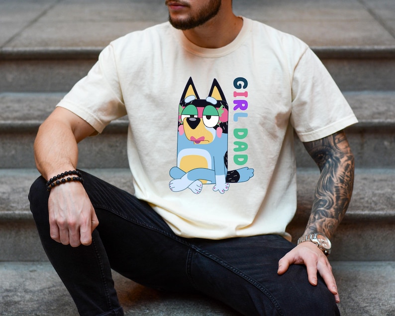 May include: A cream-colored short-sleeve t-shirt featuring a graphic of a blue cartoon dog with pink lips and green eyeshadow sitting in a cross-legged position. The text "GIRL DAD" is printed vertically in colorful letters next to the character.