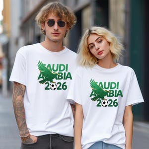 Pu&ograve; includere: T-shirt bianche con una grafica verde raffigurante un'aquila stilizzata, un pallone da calcio e la scritta "SAUDI ARABIA 2026". Le magliette sono indossate da due persone. La grafica &egrave; centrata sul davanti.