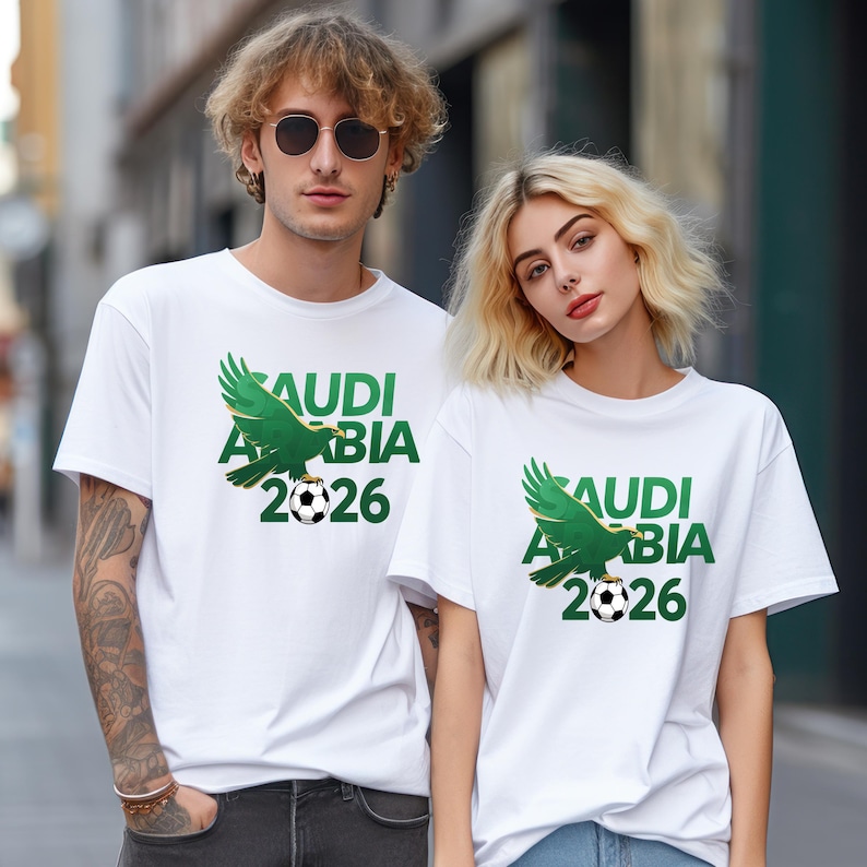 Pu&ograve; includere: T-shirt bianche con una grafica verde raffigurante un'aquila stilizzata, un pallone da calcio e la scritta "SAUDI ARABIA 2026". Le magliette sono indossate da due persone. La grafica &egrave; centrata sul davanti.