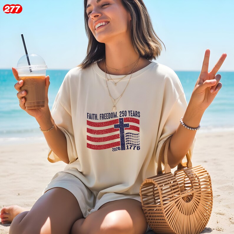 Pode incluir: Camiseta creme com um design patri&oacute;tico com uma cruz e elementos da bandeira americana. A camisa tem o texto "Faith. Freedom. 250 Years" e os anos 2026 e 1776. Uma pessoa est&aacute; vestindo a camisa e segurando uma bebida.