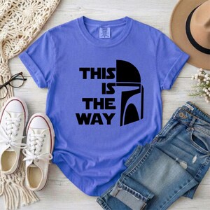 May include: A periwinkle blue t-shirt with the text "THIS IS THE WAY" in black, along with a black helmet graphic. The shirt is paired with blue jeans, white sneakers, and a brown hat.