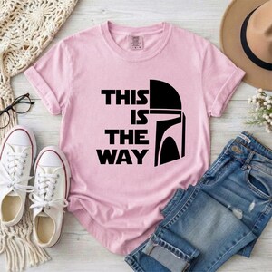 May include: A light pink t-shirt with the phrase "THIS IS THE WAY" in black block letters and a black helmet graphic. The shirt is folded, with white sneakers, blue jeans, and a hat in the background.
