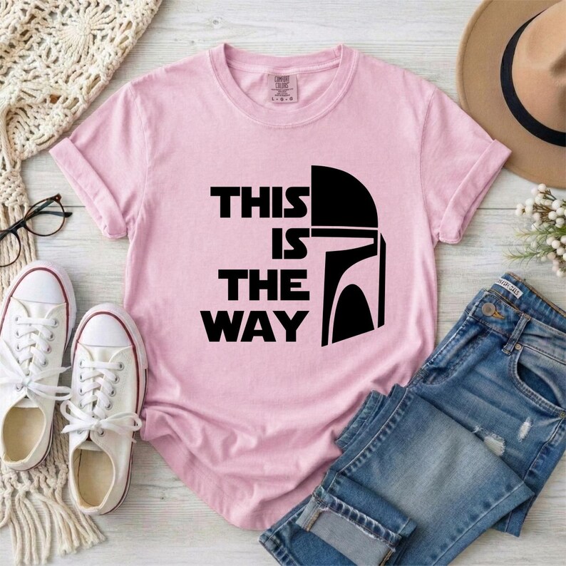 May include: A light pink t-shirt with the phrase "THIS IS THE WAY" in black block letters and a black helmet graphic. The shirt is folded, with white sneakers, blue jeans, and a hat in the background.