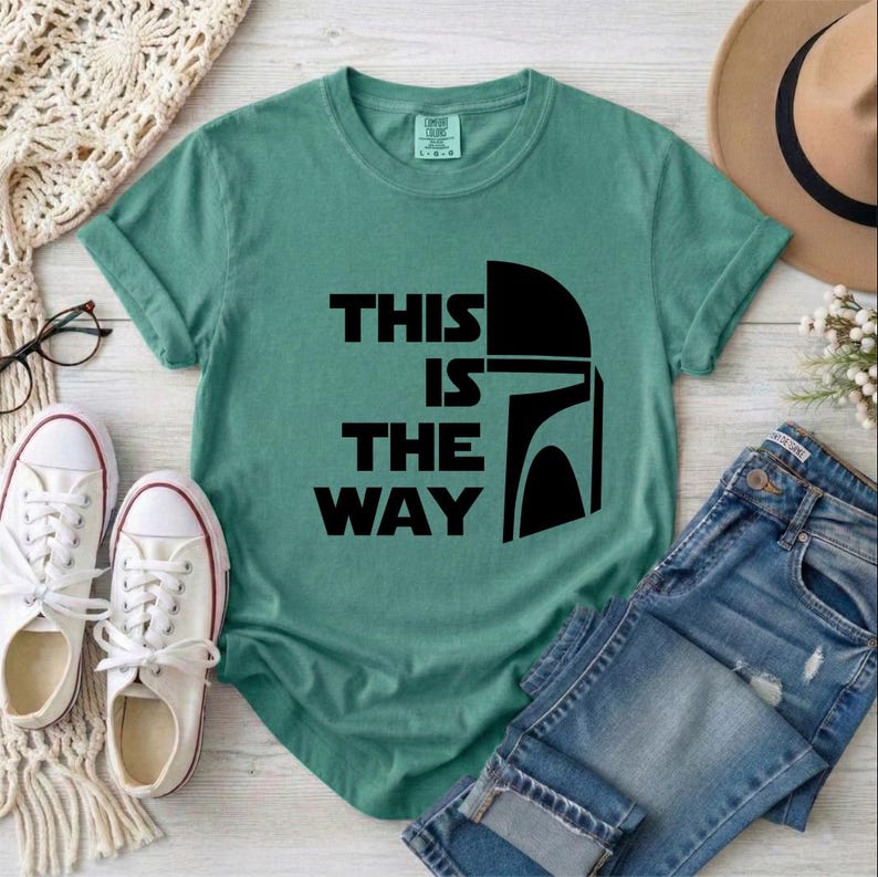 May include: A teal t-shirt with the text "THIS IS THE WAY" and a black helmet graphic. The shirt is paired with blue jeans, white sneakers, and a tan hat. The shirt is a casual style.