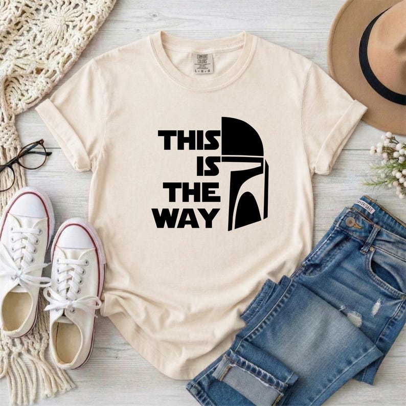 May include: Cream-colored t-shirt with the phrase "THIS IS THE WAY" in black block letters and a black helmet graphic. The shirt is paired with blue jeans, white sneakers, and a brown hat.