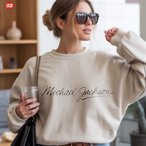 May include: A beige sweatshirt with the text "Michael Jackson" in a cursive font. The sweatshirt has long sleeves and a crew neck. The image shows a person wearing sunglasses and holding a clear cup.